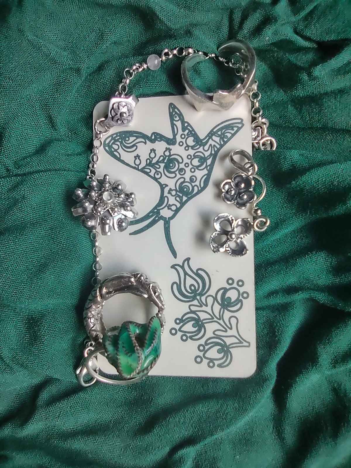 A business card front with a moth and a flower drawing with various pieces of jewelry around it on a green piece of fabric